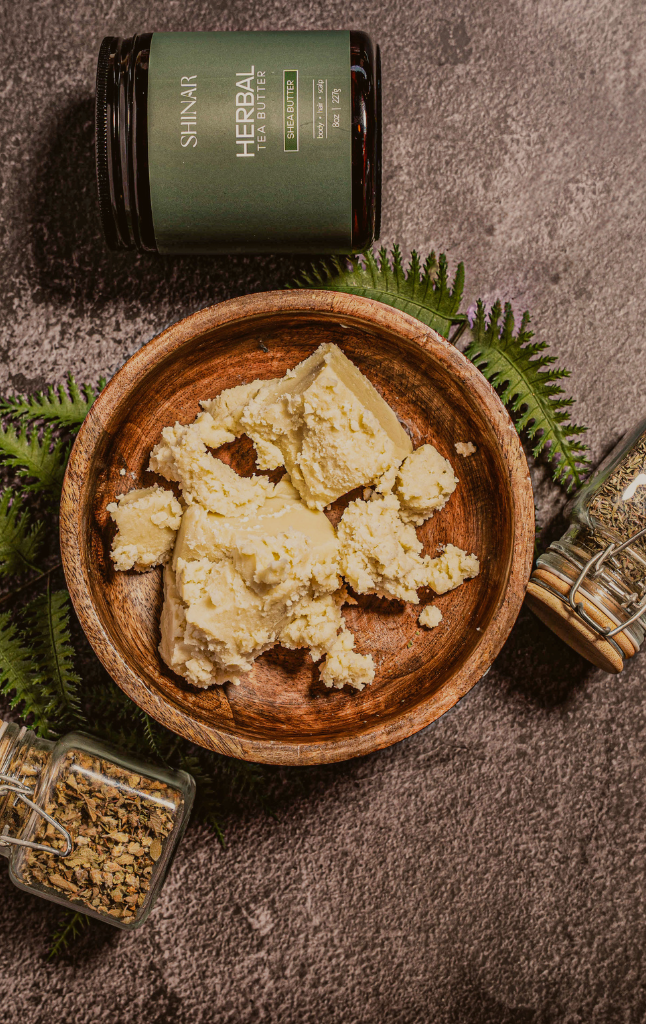 Wooden bowl containing raw shea butter, tea butter jar labeled 'Herbal Shinar', and green leaves on a dark surface